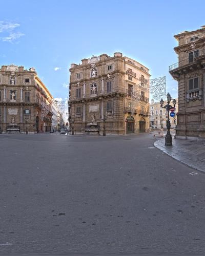 A 360-degree panoramic photograph of an octagonal city square featuring four ornate, curved corner buildings with statues and fountains, under a blue sky with some construction scaffolding visible.