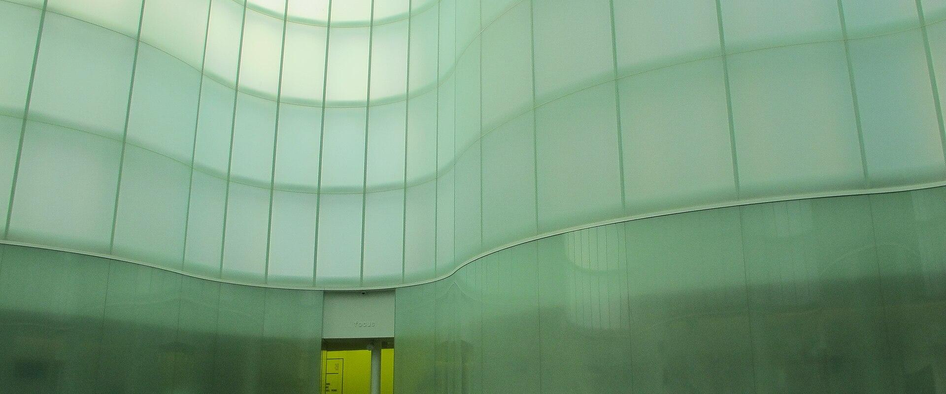 The image shows the interior of a modern, circular atrium with curved walls made of translucent green-tinted panels and a glowing yellow-lit doorway at the base; signage reading “focus” is visible above the doorway.