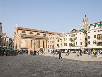 The image shows a large, open public square paved with stone, surrounded by historic buildings including a prominent brick church with arched windows and a bell tower, with people walking and sitting at outdoor cafes; signage or identifying text i