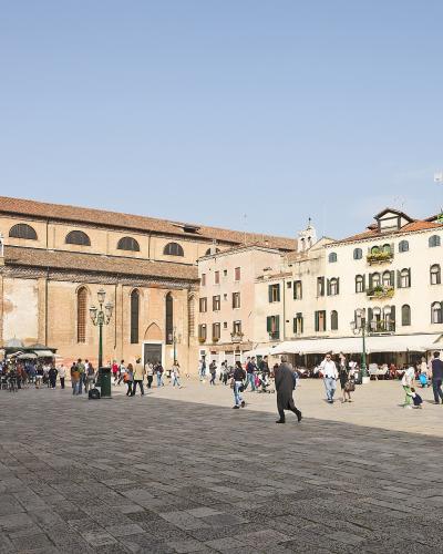 The image shows a large, open public square paved with stone, surrounded by historic buildings including a prominent brick church with arched windows and a bell tower, with people walking and sitting at outdoor cafes; signage or identifying text i