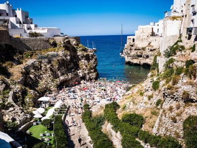 A high-angle view of a crowded pebble beach nestled between steep limestone cliffs and white-washed buildings, with boats anchored in the blue sea beyond.