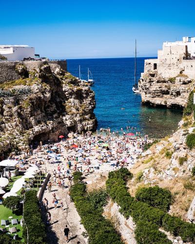 A high-angle view of a crowded pebble beach nestled between steep limestone cliffs and white-washed buildings, with boats anchored in the blue sea beyond.