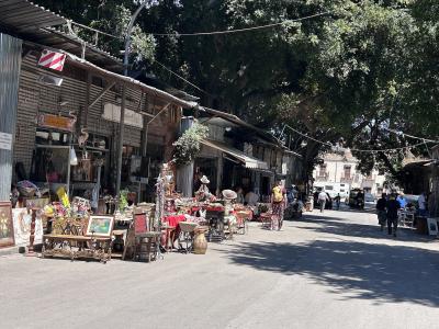 The image shows an outdoor flea market with stalls under corrugated metal roofs, displaying vintage furniture, antiques, and miscellaneous goods, with people browsing on a paved street shaded by large trees.