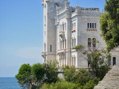 A white, Gothic Revival-style castle with a prominent clock tower, situated on a rocky cliff overlooking the sea, framed by green trees and a stone wall in the foreground.