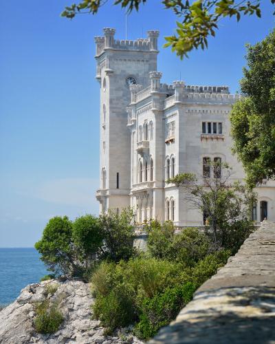 A white, Gothic Revival-style castle with a prominent clock tower, situated on a rocky cliff overlooking the sea, framed by green trees and a stone wall in the foreground.