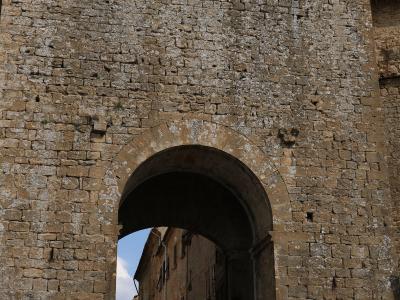 A large, medieval stone gatehouse with crenellations and a rounded archway, featuring a "no entry" traffic sign and a yellow 'S' symbol on the lower right wall, looking out onto a narrow street with pedestrians.