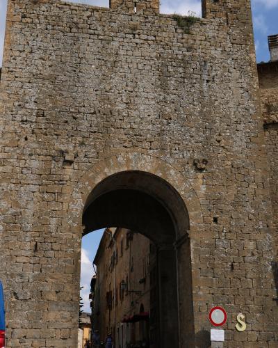 A large, medieval stone gatehouse with crenellations and a rounded archway, featuring a "no entry" traffic sign and a yellow 'S' symbol on the lower right wall, looking out onto a narrow street with pedestrians.