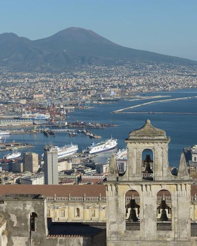 A high-angle panoramic view of the city of Naples, Italy, featuring the port and harbor in the mid-ground with Mount Vesuvius dominating the background, and the bell tower and roofline of the Certosa di San Martino (Abbey of San Martino) in the im