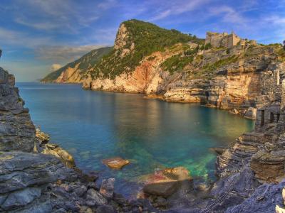 A scenic coastal view from within a rocky sea cave looking out toward a sunlit cliffside with a fortress and green hill, featuring layered limestone rock formations framing the turquoise water.
