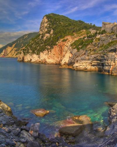 A scenic coastal view from within a rocky sea cave looking out toward a sunlit cliffside with a fortress and green hill, featuring layered limestone rock formations framing the turquoise water.