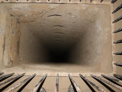 A downward-looking view through a square metal grate into a deep, narrow, rectangular shaft carved into light-colored tufa rock, with a person's feet visible on the left edge.