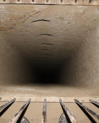 A downward-looking view through a square metal grate into a deep, narrow, rectangular shaft carved into light-colored tufa rock, with a person's feet visible on the left edge.