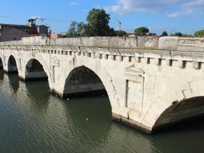 A stone Roman arch bridge with multiple arches spanning a river, featuring decorative corbels along the parapet and visible weathering on the stonework, with buildings and construction cranes in the background under a blue sky.