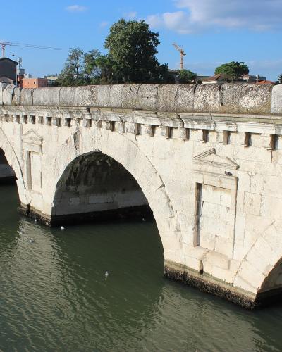 A stone Roman arch bridge with multiple arches spanning a river, featuring decorative corbels along the parapet and visible weathering on the stonework, with buildings and construction cranes in the background under a blue sky.