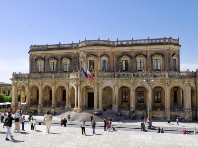 A large, ornate Baroque-style palace with a ground-floor loggia of arches and an upper story with pilasters and a balustrade, flying Italian and EU flags, situated in a sunlit public square with tourists.