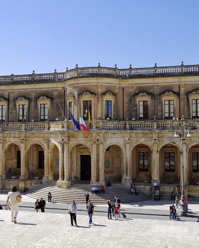 A large, ornate Baroque-style palace with a ground-floor loggia of arches and an upper story with pilasters and a balustrade, flying Italian and EU flags, situated in a sunlit public square with tourists.