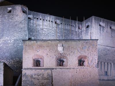 A night-time photograph of a large, illuminated stone fortress featuring prominent brickwork, arched cannon emplacements, and a central coat of arms above the cannons.