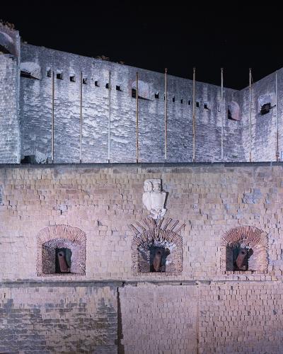 A night-time photograph of a large, illuminated stone fortress featuring prominent brickwork, arched cannon emplacements, and a central coat of arms above the cannons.