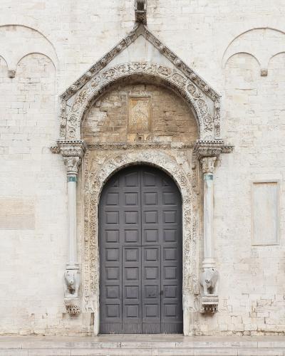 The image shows a close-up of an ornate, Gothic-style stone portal with a pointed arch, intricate carvings, and two sculpted lions at the base of the columns, set into a light-colored stone facade.