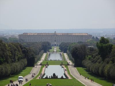 A wide-angle view of the Royal Palace of Caserta with its long, symmetrical gardens, reflecting pools, and fountains leading up to the palace facade.
