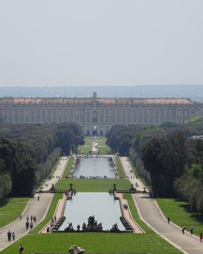 A wide-angle view of the Royal Palace of Caserta with its long, symmetrical gardens, reflecting pools, and fountains leading up to the palace facade.