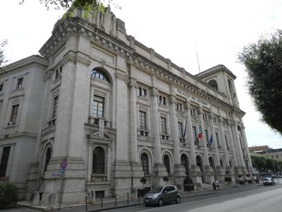 A large, classical-style stone government building with the inscription "PALAZZO DEL GOVERNO" on its facade, featuring columns, arched windows, and Italian flags, situated on a street corner with a car and pedestrians.