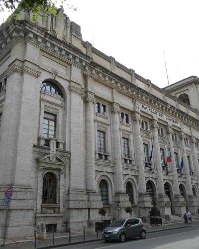 A large, classical-style stone government building with the inscription "PALAZZO DEL GOVERNO" on its facade, featuring columns, arched windows, and Italian flags, situated on a street corner with a car and pedestrians.