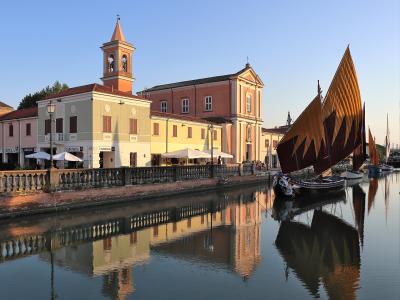 A view of a canal lined with colorful buildings and a church with a bell tower, featuring traditional sailing boats with large triangular sails moored in the water.