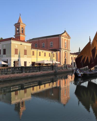 A view of a canal lined with colorful buildings and a church with a bell tower, featuring traditional sailing boats with large triangular sails moored in the water.