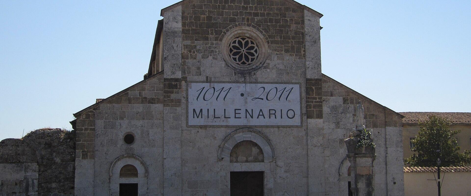 A stone church facade with a cross on top, a rose window, and a large banner reading "1011 · 2011 MILLENARIO"; a person on a motorcycle is visible to the right.