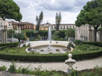 A view of Villa Bellini park in Catania, featuring a central fountain, manicured hedges, statues, and surrounding historic buildings under a cloudy sky.