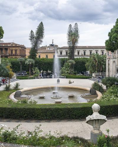 A view of Villa Bellini park in Catania, featuring a central fountain, manicured hedges, statues, and surrounding historic buildings under a cloudy sky.