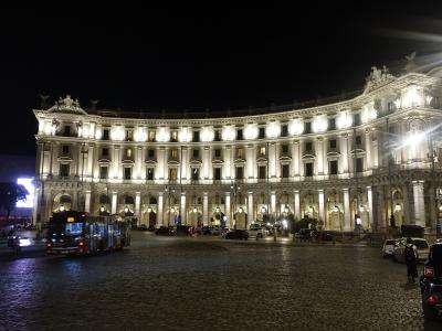 A wide-angle view of a large urban square featuring a central fountain with sculptural groups, surrounded by historic brick ruins and modern buildings under a cloudy sky.