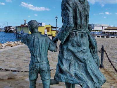 A bronze statue of a woman and two children looking out at a harbor, with a yellow building and waterfront structures in the background.