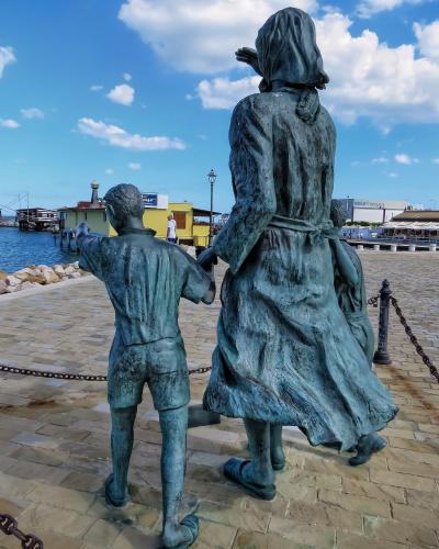 A bronze statue of a woman and two children looking out at a harbor, with a yellow building and waterfront structures in the background.