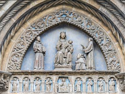 A close-up of an ornate Gothic stone tympanum and archway featuring a central sculpture of the Madonna and Child flanked by saints, with a row of smaller figures in niches below, set against a blue background.