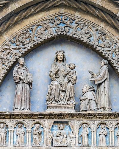 A close-up of an ornate Gothic stone tympanum and archway featuring a central sculpture of the Madonna and Child flanked by saints, with a row of smaller figures in niches below, set against a blue background.