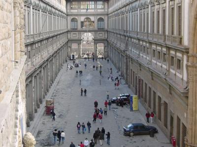 A close-up view of two large wooden doors set into a stone building facade, labeled "REALI" and "POSTE", with a construction hoarding in the foreground displaying an image of the Uffizi Gallery facade and its name.