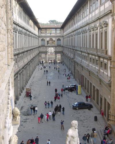 A close-up view of two large wooden doors set into a stone building facade, labeled "REALI" and "POSTE", with a construction hoarding in the foreground displaying an image of the Uffizi Gallery facade and its name.
