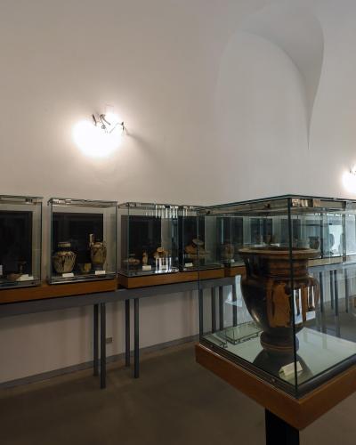 A museum gallery interior featuring glass display cases containing ancient pottery and artifacts, with white walls and arched ceilings.