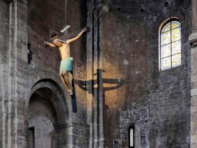 The image shows a large, suspended crucifix sculpture inside a stone church interior, with a stained-glass window and arched architecture visible in the background.