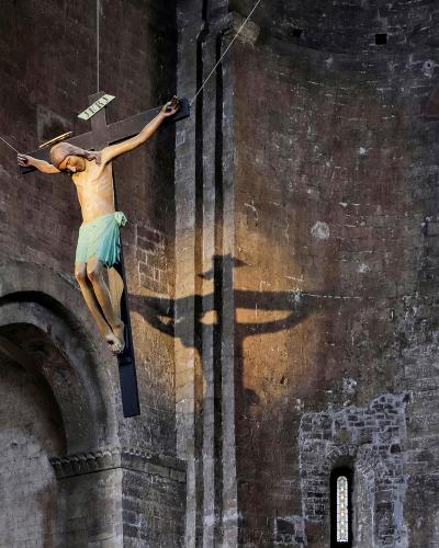 The image shows a large, suspended crucifix sculpture inside a stone church interior, with a stained-glass window and arched architecture visible in the background.