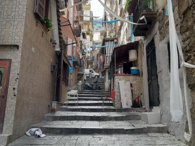 A narrow, steep stone-paved alleyway with stairs ascending between old residential buildings, featuring laundry lines strung overhead, balconies with plants, and a parked scooter at the top.