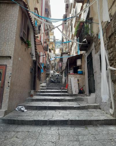 A narrow, steep stone-paved alleyway with stairs ascending between old residential buildings, featuring laundry lines strung overhead, balconies with plants, and a parked scooter at the top.