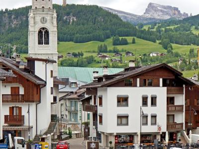 A street-level view of a European alpine town featuring a prominent white church tower with a green spire, traditional chalet-style buildings, parked cars, and a backdrop of green hills and rocky mountain peaks.