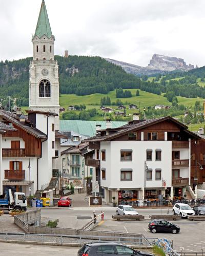 A street-level view of a European alpine town featuring a prominent white church tower with a green spire, traditional chalet-style buildings, parked cars, and a backdrop of green hills and rocky mountain peaks.