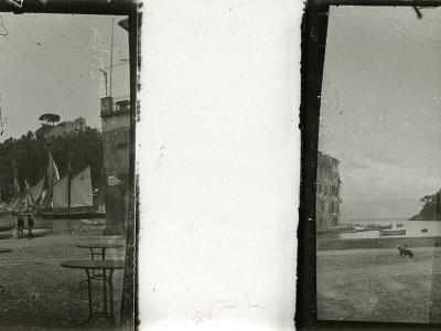 A vintage black-and-white stereoscopic photograph showing a cobblestone waterfront square with outdoor cafe tables, several traditional sailboats moored in the harbor, and a hillside village with buildings and trees rising behind the water.