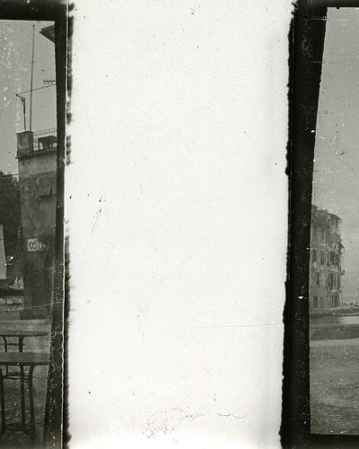 A vintage black-and-white stereoscopic photograph showing a cobblestone waterfront square with outdoor cafe tables, several traditional sailboats moored in the harbor, and a hillside village with buildings and trees rising behind the water.