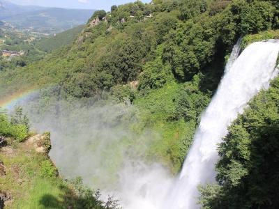 A large waterfall cascading down a steep, forested cliff face with a visible rainbow in the mist below and a valley landscape in the background.