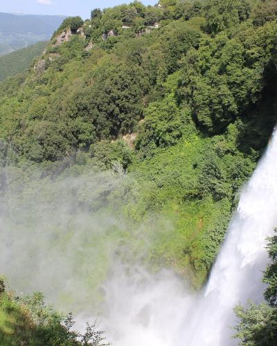 A large waterfall cascading down a steep, forested cliff face with a visible rainbow in the mist below and a valley landscape in the background.